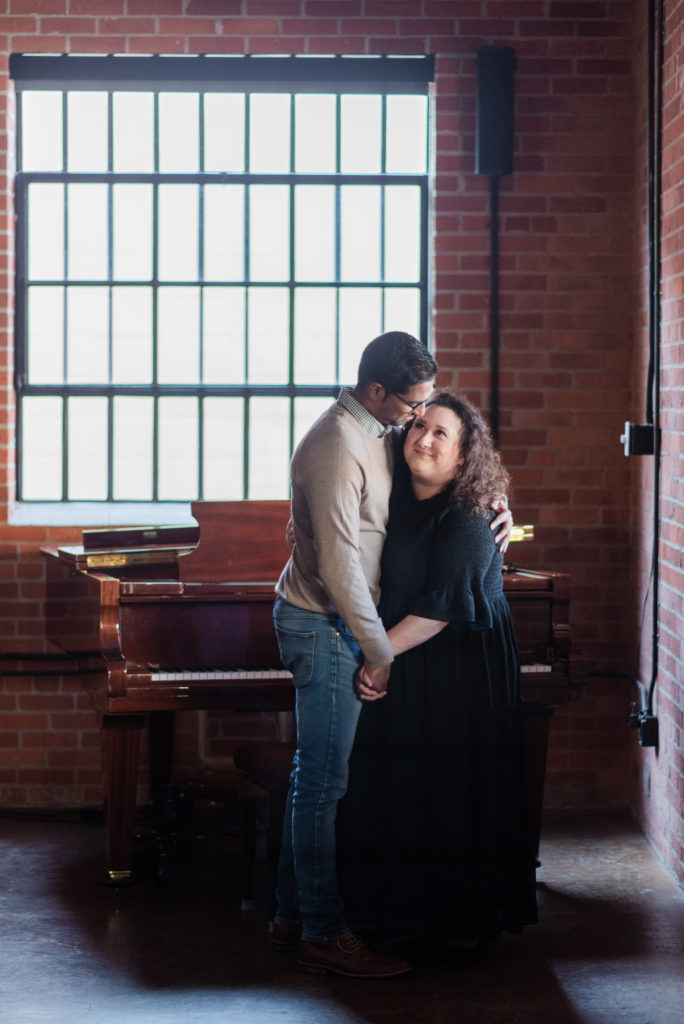 A couple embraces in front of a grand piano and a window. Downtown Greer Portrait Sessions at Randall House