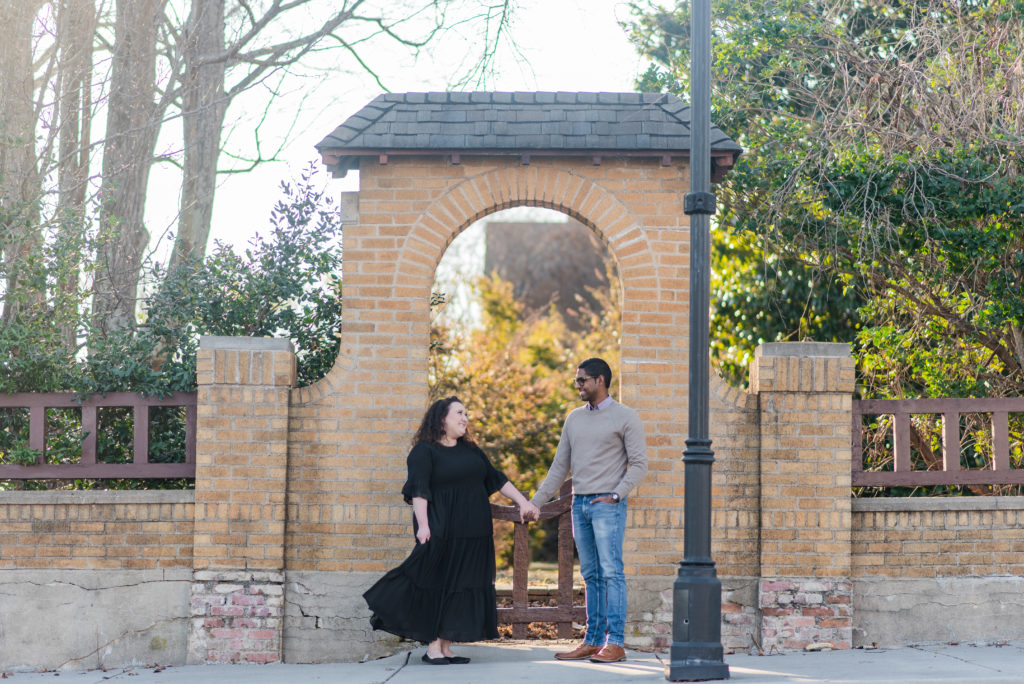 A couple dances outside in front of an orange brick arch with greenery and a lamp post. Downtown Greer Portrait Sessions at Randall House