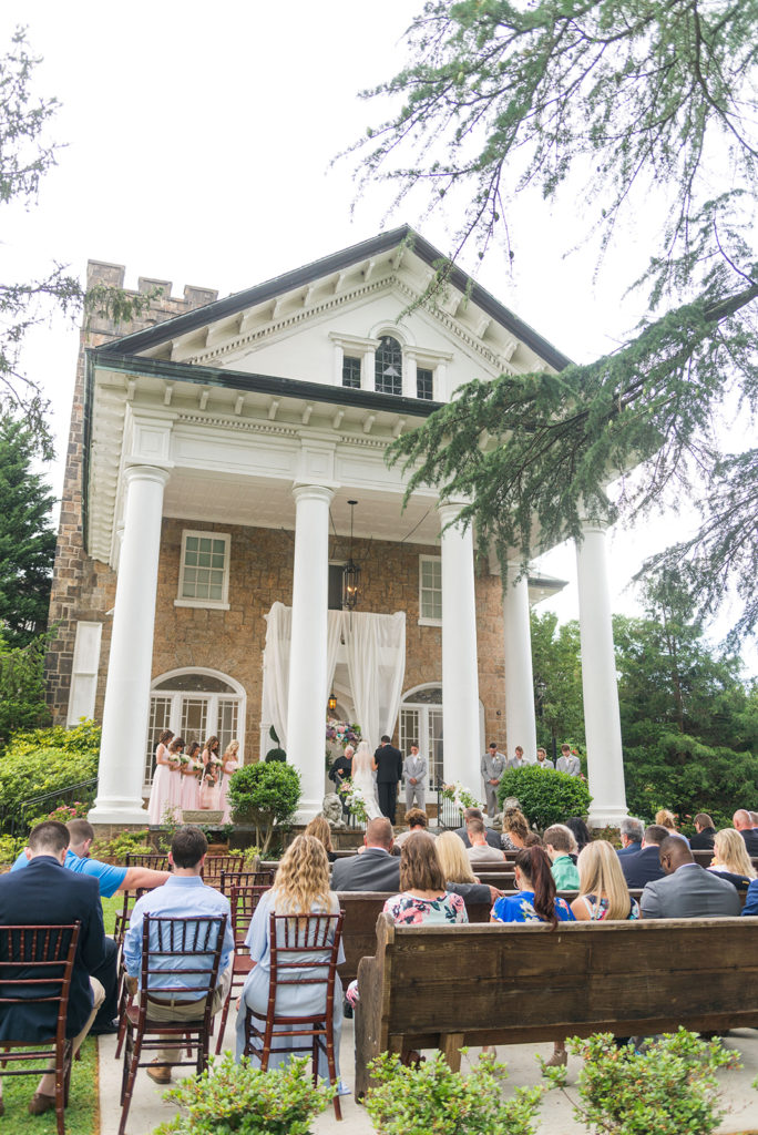 ceremony at the gassaway mansion. it is a large house with tall, white columns. The guests are sitting outside in wooden chairs and pews. The ceremony is taking place on the steps.