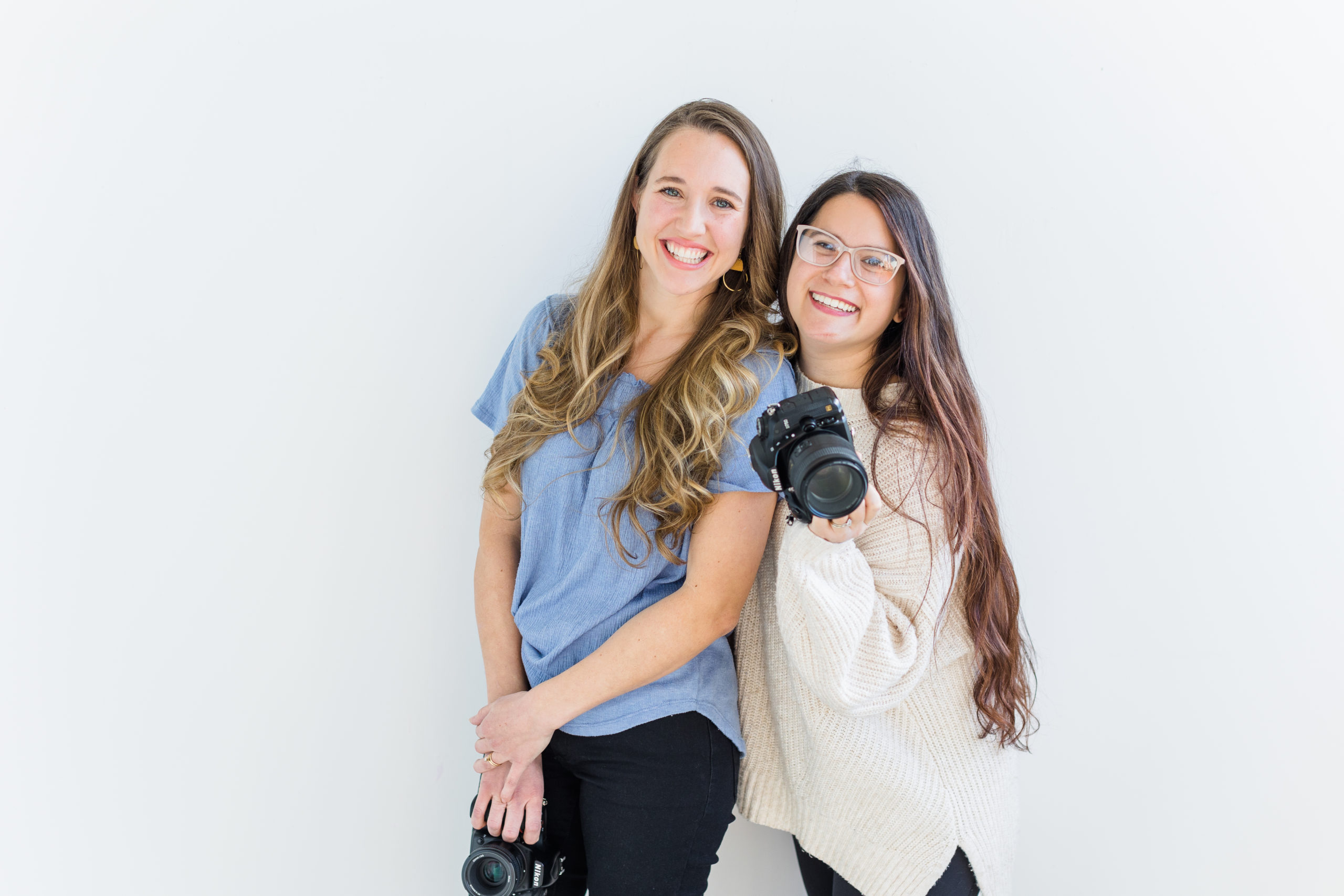 stephanie is wearing a blue shirt and black pants. christi is wearing a white sweater and black leggings. they are both smiling at the camera and christi is holding her camera