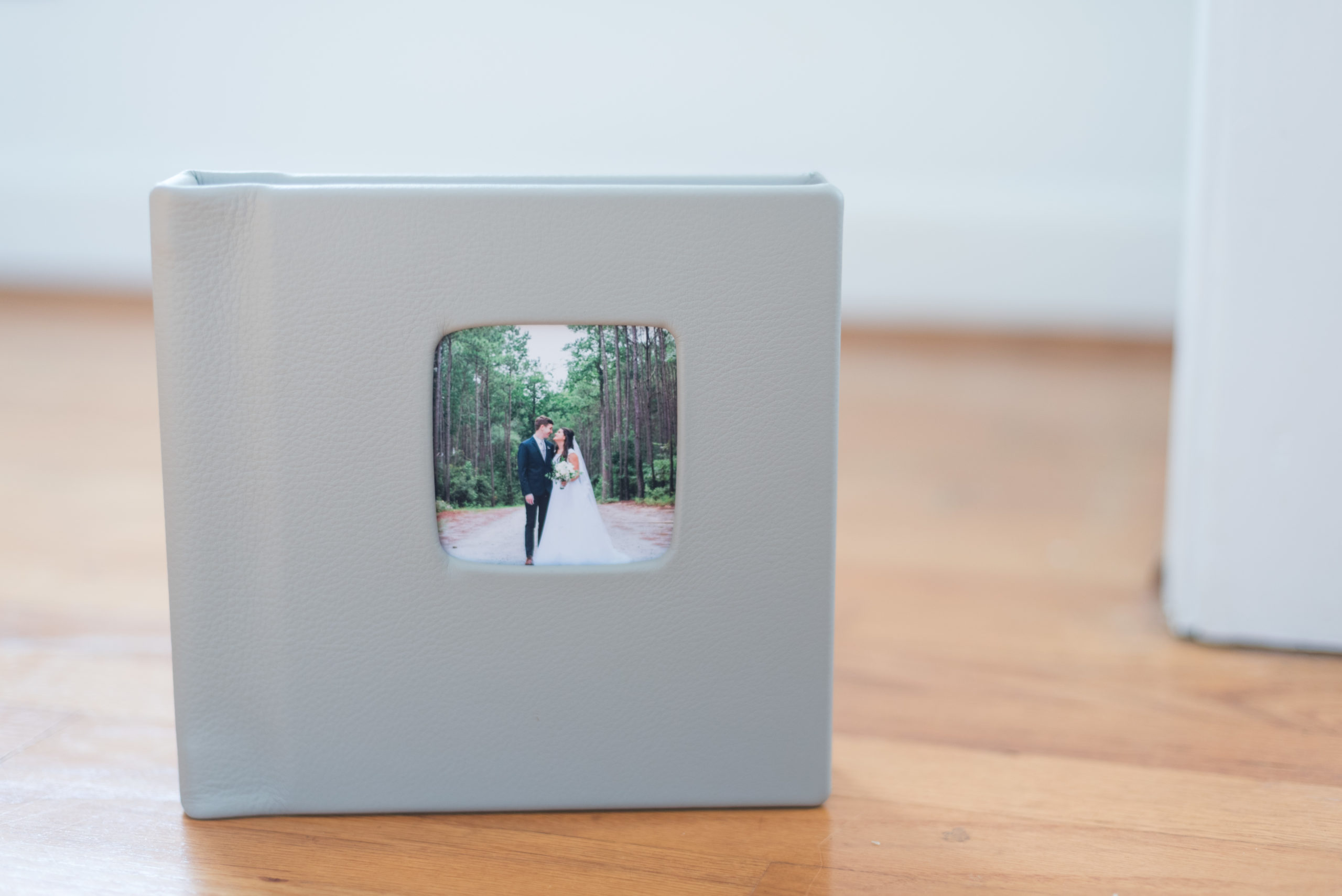 A gray, leather-bound wedding album with a cutout on the front showcasing a wedding photo on the front