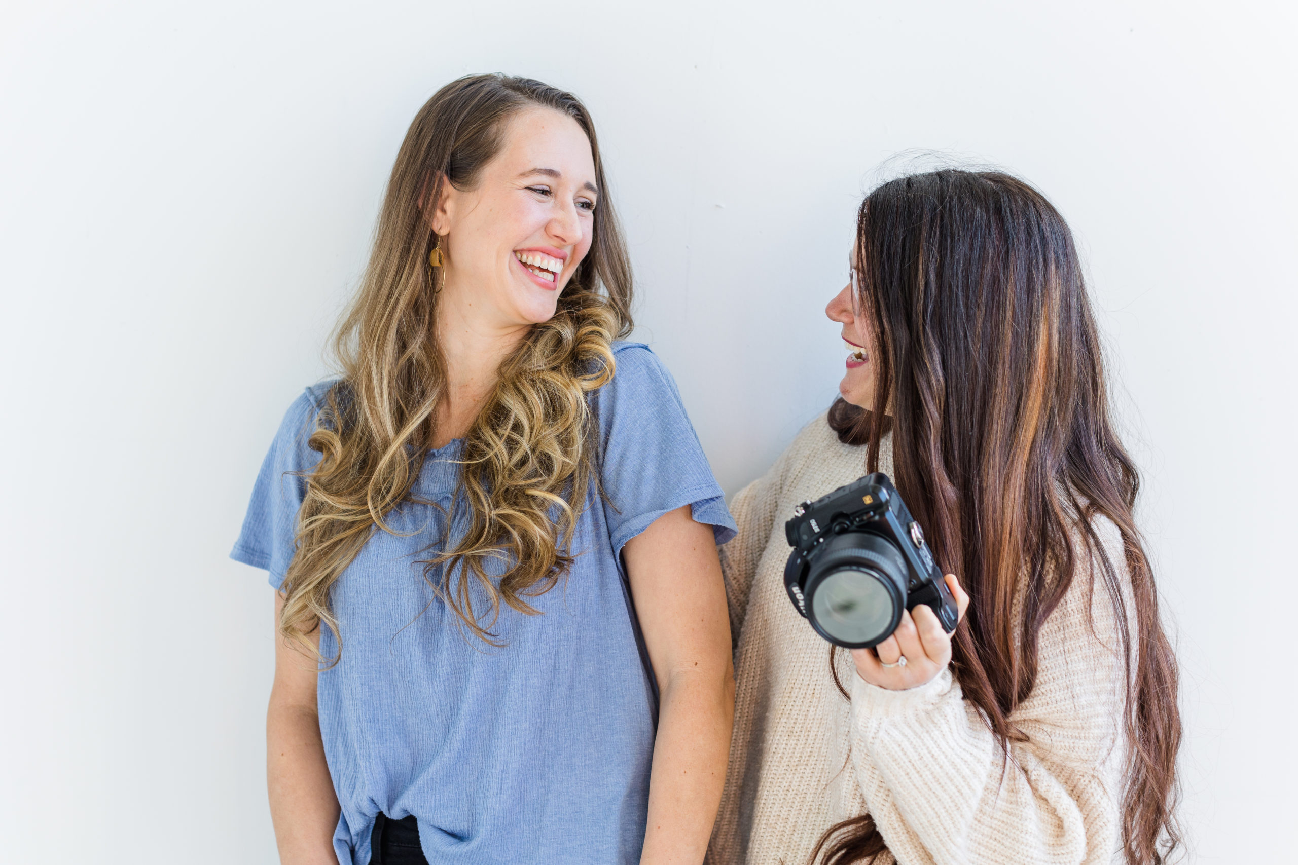 Stephanie is wearing a blue shirt and black pants. Christi is beside her wearing a white oversized sweater and black pants. They are both holding their cameras and laughing at each other. Top 5 Questions to Ask Your Photographer