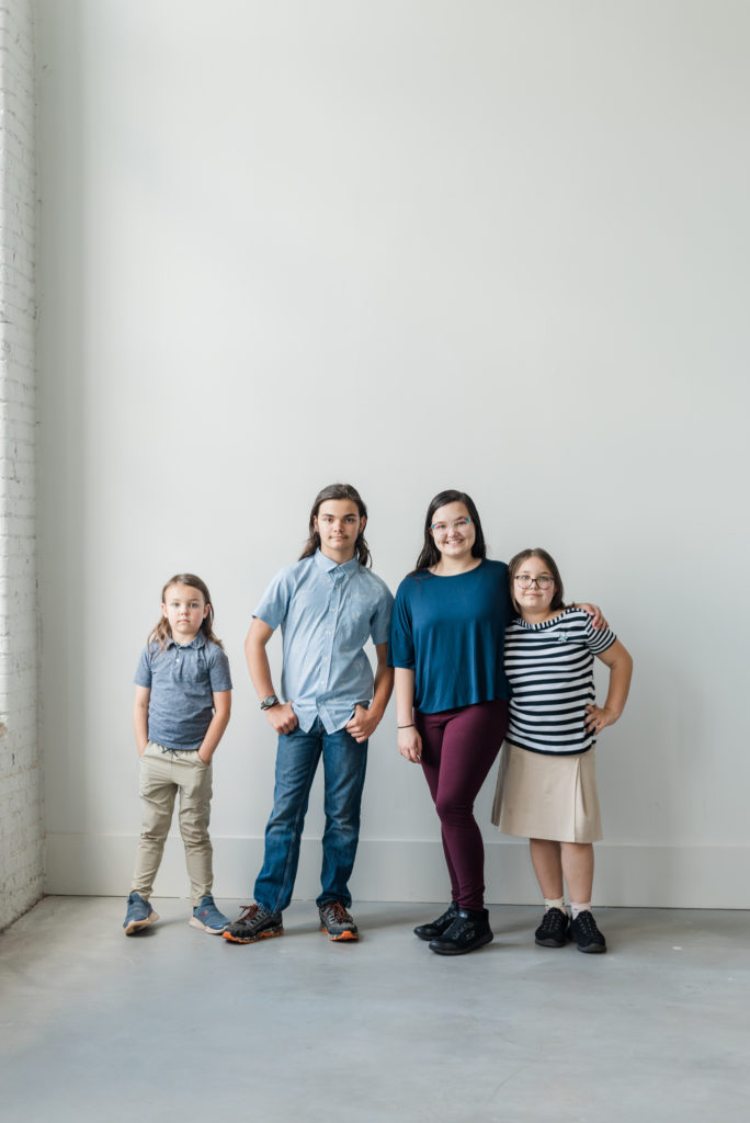 Greenville Family Photos at Cultivate Studios - A group of siblings are smiling at the camera standing in front of a white wall. It is a teenaged girl, teenaged boy, grade-school girl, and grade-school boy. 