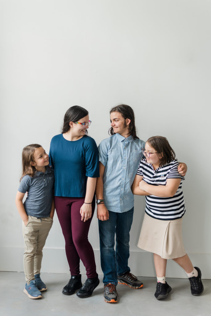 Greenville Family Photos at Cultivate Studios - A group of siblings are laughing at each other standing in front of a white wall. It is a teenaged girl, teenaged boy, grade-school girl, and grade-school boy. 