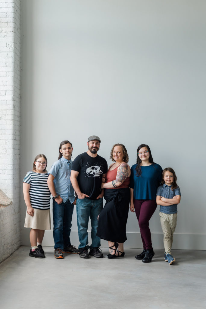 Greenville Family Photos at Cultivate Studios - A family is smiling at the camera in front of a white wall. It is a mom, dad, teenaged girl, teenaged boy, grade-school girl, and grade-school boy. 
