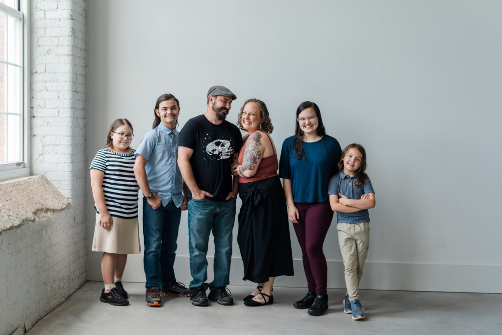 Greenville Family Photos at Cultivate Studios - A family is laughing at the camera in front of a white wall. It is a mom, dad, teenaged girl, teenaged boy, grade-school girl, and grade-school boy. 