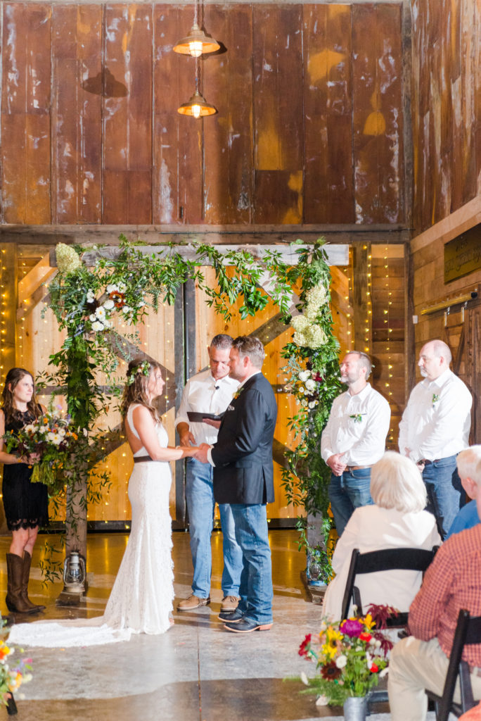 Iron Oak Barn Wedding in Pendleton SC - A couple gets married in front of a flowered wooden arbor in a rustic barn