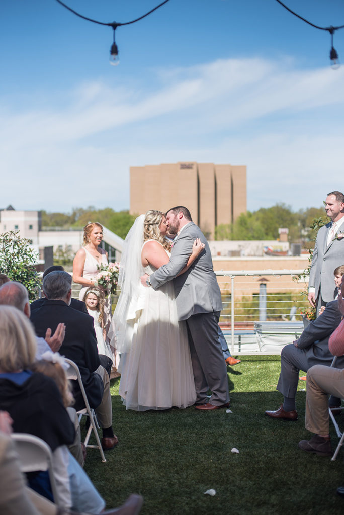 A bride and groom share a kiss at a wedding ceremony at The Upper Room in Greenville, SC on their rooftop in front of the Greenville skyline