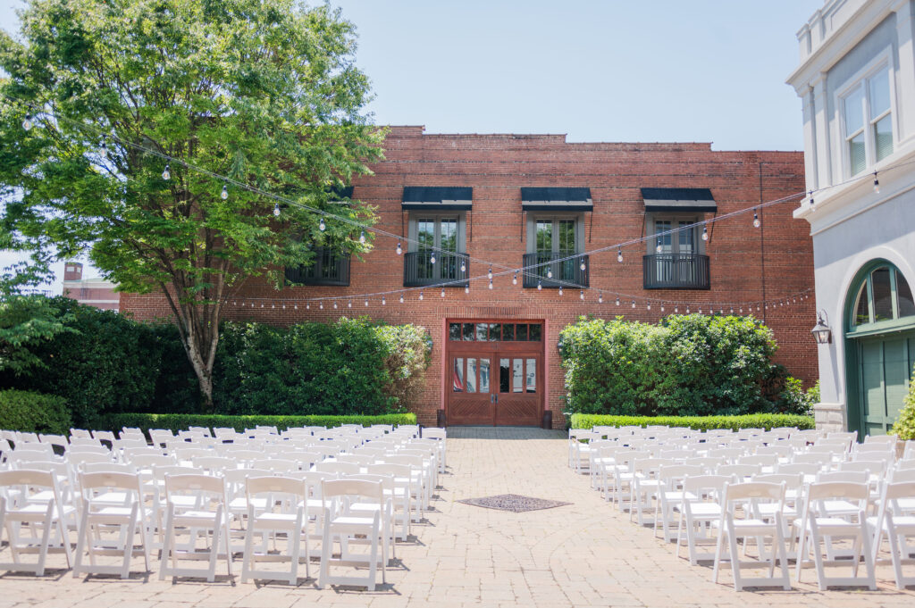 The ceremony space at the wedding venue in The Bleckley Inn. There are white chairs, a brick building with large doors, and twinkle lights intersecting above the chairs.