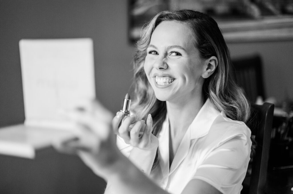 A bride is smiling while putting on her lipstick lit by natural light in the bride's suite at The Bleckley Inn